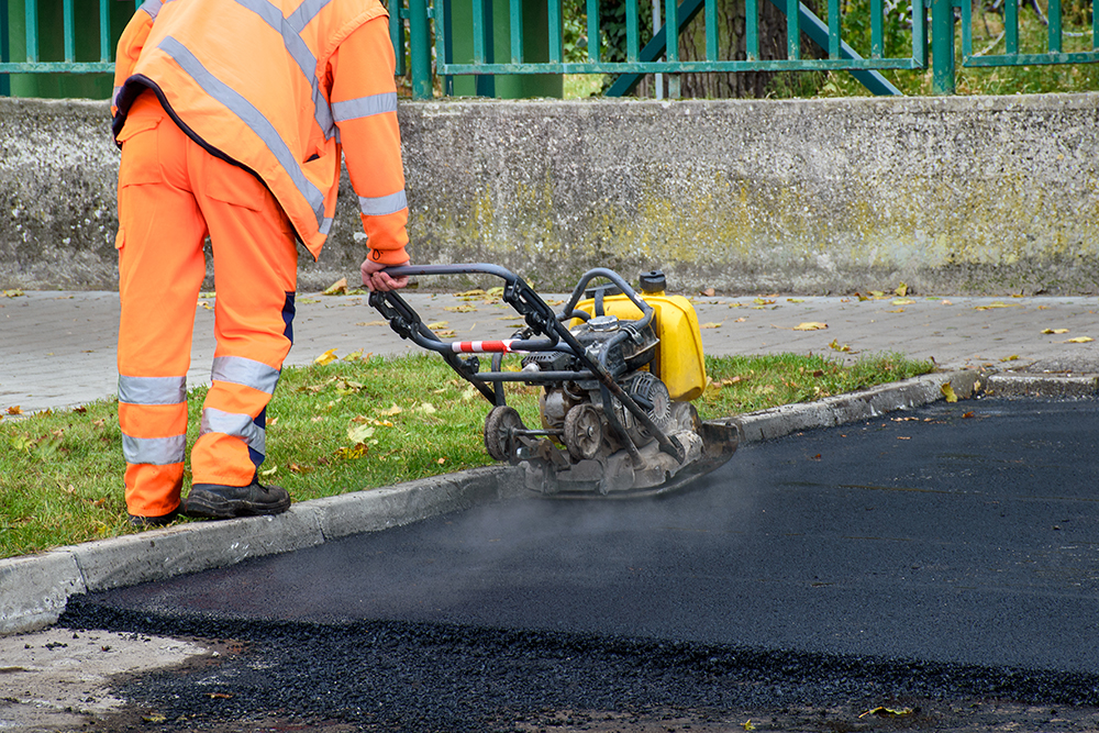 Asphalt Maintenance Paving worker uses vibratory plate compactor to compact new asphalt near curbstones